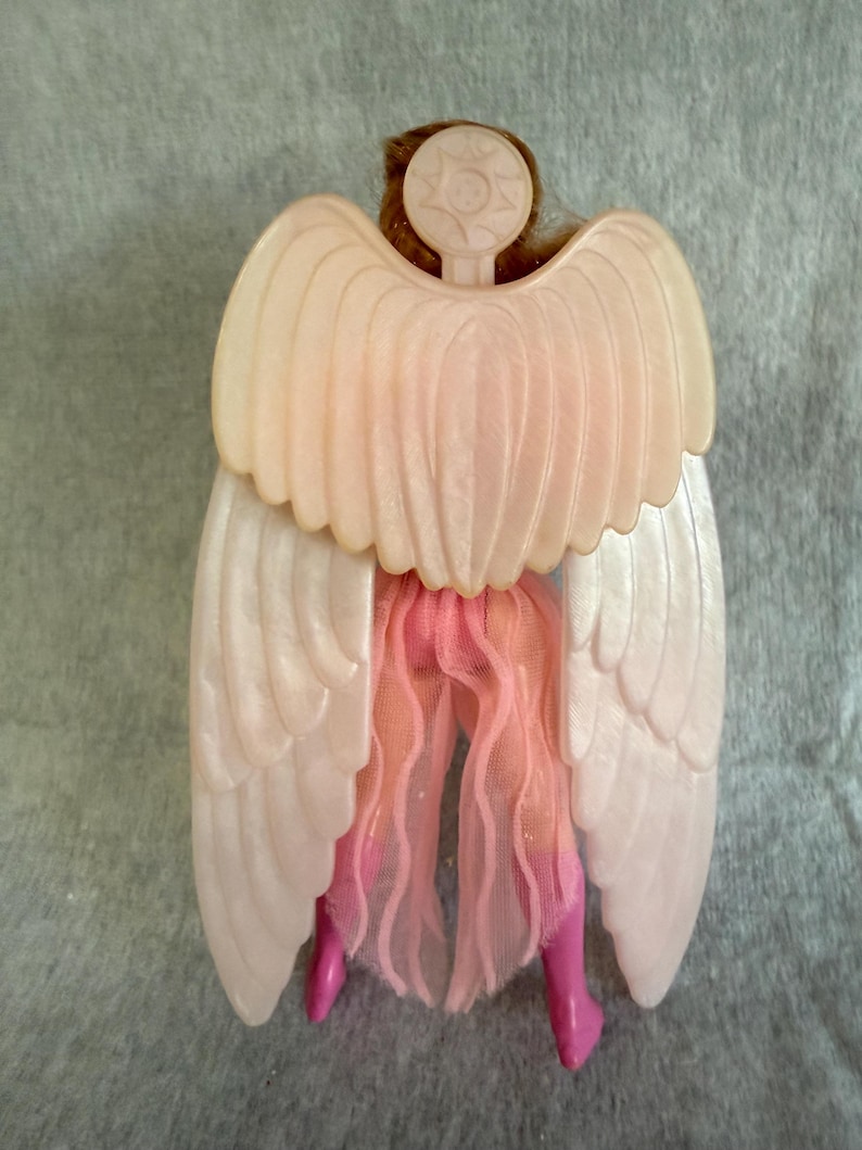 May include: A doll figure viewed from the back, featuring large, pale, pearlescent plastic wings attached to a circular emblem at the neck. The doll wears a sheer, light pink fabric skirt and bright pink boots, with brown hair visible above the wings.