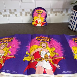 May include: A purple fabric featuring a repeating graphic of Princess Power with long blonde hair, a white dress, and a gold sword. The design includes a bright pink starburst background and the text Princess Power printed in yellow letters.