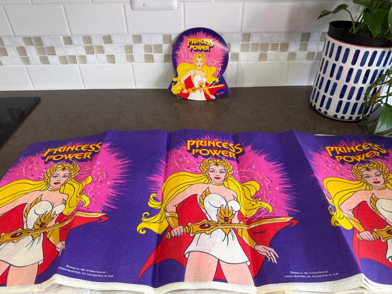 May include: A purple fabric featuring a repeating graphic of Princess Power with long blonde hair, a white dress, and a gold sword. The design includes a bright pink starburst background and the text Princess Power printed in yellow letters.