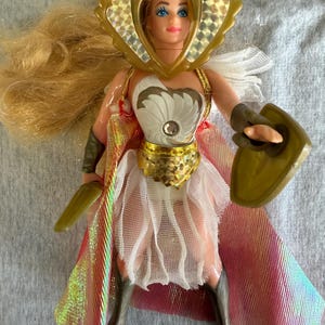 May include: A blonde doll wearing a gold-colored headpiece with holographic accents, a white bodice with a central gem, a gold-colored waist belt, and a white skirt. The doll features a long iridescent cape, dark-colored boots, and holds a shield.