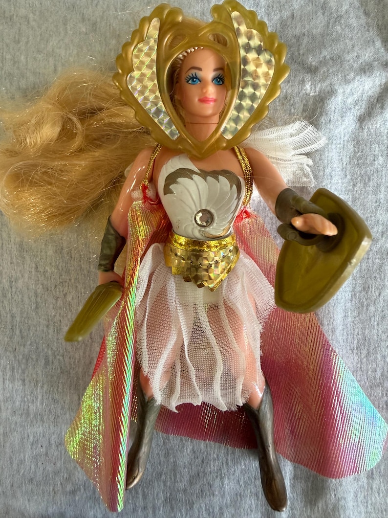 May include: A blonde doll wearing a gold-colored headpiece with holographic accents, a white bodice with a central gem, a gold-colored waist belt, and a white skirt. The doll features a long iridescent cape, dark-colored boots, and holds a shield.
