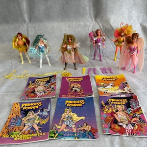 May include: A collection of six Princess of Power action figures with various accessories and six illustrated storybooks. The dolls feature colorful outfits, wings, and hair, while the books display titles like The Hidden Symbols Mystery.