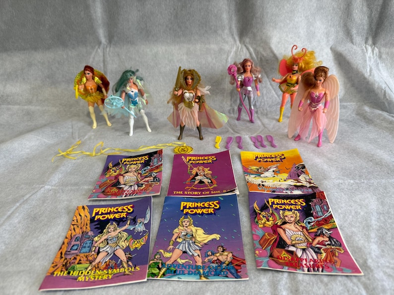 May include: A collection of six Princess of Power action figures with various accessories and six illustrated storybooks. The dolls feature colorful outfits, wings, and hair, while the books display titles like The Hidden Symbols Mystery.