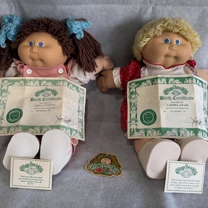 May include: Two Cabbage Patch Kids dolls with birth certificates. One doll has brown yarn hair with blue ribbons and a pink outfit. The other has blonde yarn hair and a red dress. Both have white shoes and include small informational cards and a logo sticker.