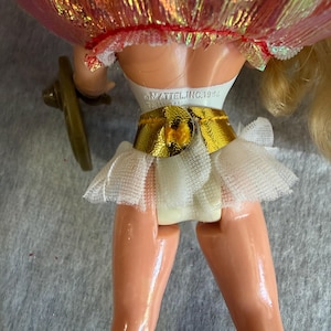 May include: A doll with blonde hair wearing a white bodice, a metallic gold belt, and a white tulle skirt. The back of the doll features the text MATTEL, INC. 1994 TAIWAN embossed on the white plastic torso. A small brown accessory is attached to the side.