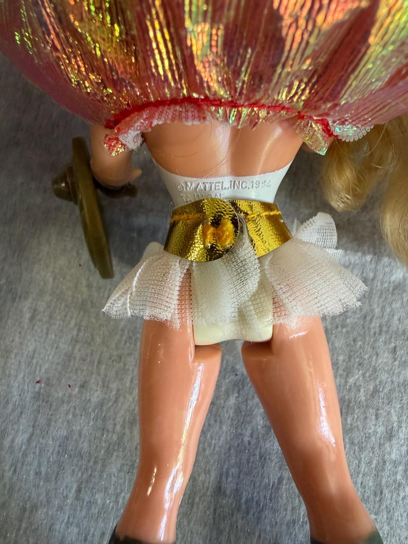 May include: A doll with blonde hair wearing a white bodice, a metallic gold belt, and a white tulle skirt. The back of the doll features the text MATTEL, INC. 1994 TAIWAN embossed on the white plastic torso. A small brown accessory is attached to the side.