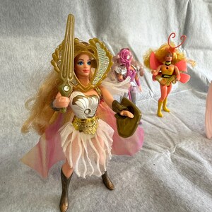 May include: A blonde action figure wearing a gold headpiece, white bodice with a silver accent, and a white layered skirt with a pink cape. The figure holds a gold sword and shield. Two smaller figures stand in the background, one purple and one yellow.