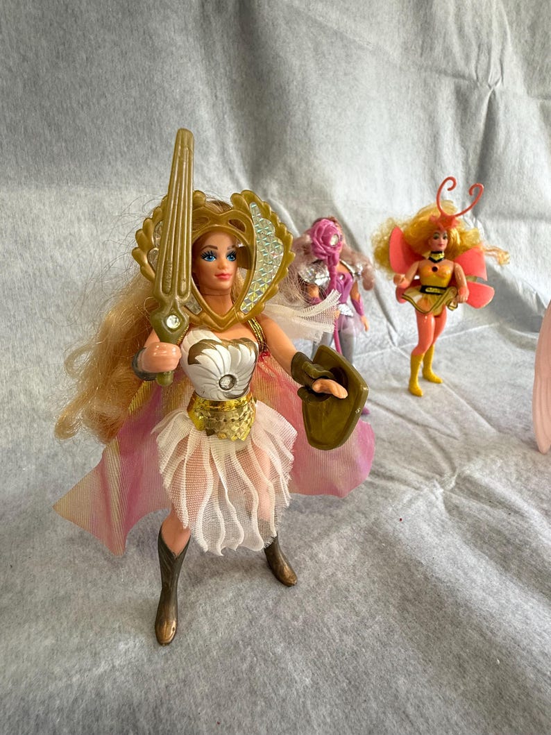 May include: A blonde action figure wearing a gold headpiece, white bodice with a silver accent, and a white layered skirt with a pink cape. The figure holds a gold sword and shield. Two smaller figures stand in the background, one purple and one yellow.