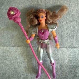May include: A plastic action figure with long wavy light brown hair, wearing a purple bodice with a silver chest accent, silver shoulder pads, silver leggings, and purple boots. The figure holds a long pink staff topped with a pink rose and a gem.