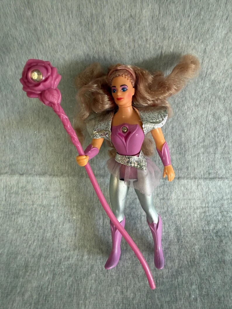 May include: A plastic action figure with long wavy light brown hair, wearing a purple bodice with a silver chest accent, silver shoulder pads, silver leggings, and purple boots. The figure holds a long pink staff topped with a pink rose and a gem.