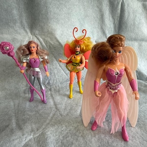 May include: Three dolls with distinct outfits and accessories. The left doll has wavy hair, a purple and silver outfit, and a pink staff. The center doll has yellow hair, orange antennae, and a yellow and orange outfit. The right doll has large wings.