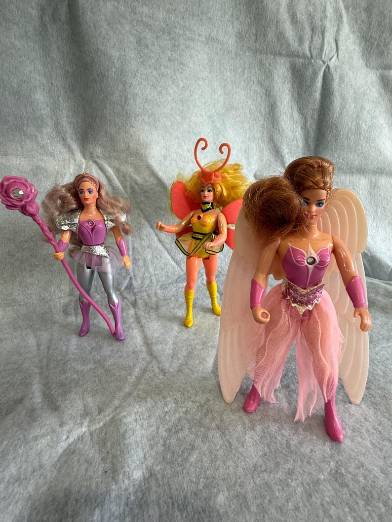 May include: Three dolls with distinct outfits and accessories. The left doll has wavy hair, a purple and silver outfit, and a pink staff. The center doll has yellow hair, orange antennae, and a yellow and orange outfit. The right doll has large wings.