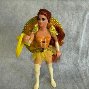 May include: A plastic action figure with long auburn hair, wearing an orange bodice with white lightning bolt accents, a gold metallic shoulder piece, a yellow tulle skirt, and white knee-high boots. The figure holds a yellow shield and circular disc.