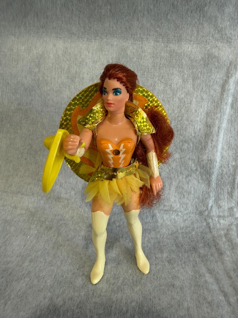 May include: A plastic action figure with long auburn hair, wearing an orange bodice with white lightning bolt accents, a gold metallic shoulder piece, a yellow tulle skirt, and white knee-high boots. The figure holds a yellow shield and circular disc.