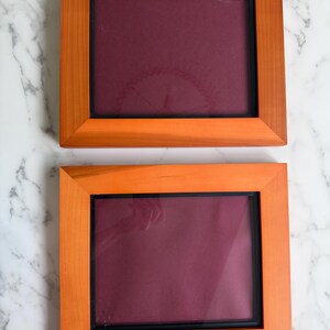 May include: Two rectangular picture frames with light brown wooden borders and a dark maroon velvet-like backing. Each frame features a thin black inner border surrounding the central display area, providing a clean and simple presentation.