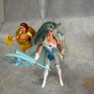 May include: Two plastic dolls. The foreground doll has long teal hair, a blue and silver bodice with a star emblem, a sheer iridescent cape, and white boots, holding a light blue circular staff. The background doll has red hair and a yellow outfit.