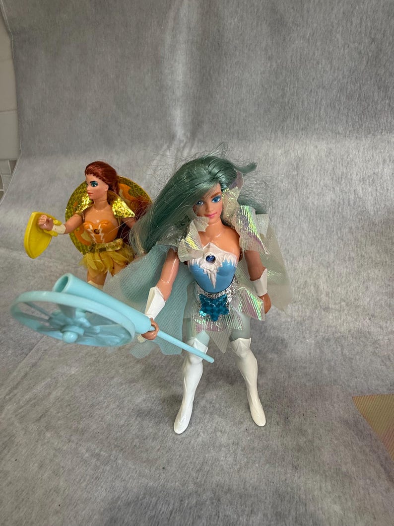 May include: Two plastic dolls. The foreground doll has long teal hair, a blue and silver bodice with a star emblem, a sheer iridescent cape, and white boots, holding a light blue circular staff. The background doll has red hair and a yellow outfit.