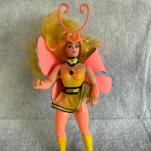 May include: A doll with long yellow hair wearing a yellow and peach outfit with large pink butterfly wings. The doll features a peach-colored headpiece with antennae, a yellow bodice with a dark center, a yellow mesh skirt, and yellow boots.