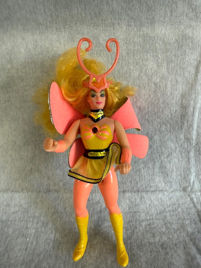 May include: A doll with long yellow hair wearing a yellow and peach outfit with large pink butterfly wings. The doll features a peach-colored headpiece with antennae, a yellow bodice with a dark center, a yellow mesh skirt, and yellow boots.