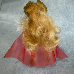 May include: A doll with long, wavy blonde hair styled with a gold-colored crown accessory. The doll is wearing a translucent, pleated red cape that flares out at the bottom, partially obscuring the lower body and legs of the figure.