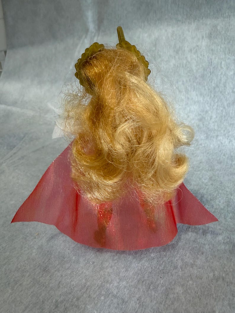 May include: A doll with long, wavy blonde hair styled with a gold-colored crown accessory. The doll is wearing a translucent, pleated red cape that flares out at the bottom, partially obscuring the lower body and legs of the figure.