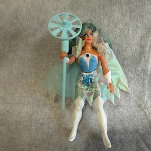 May include: A doll with teal hair wearing a blue and white bodice, a silver waist accent, and white boots. The doll features a translucent iridescent cape and holds a light blue staff topped with a circular, fan-like accessory. The figure has painted facial features and articulated limbs.