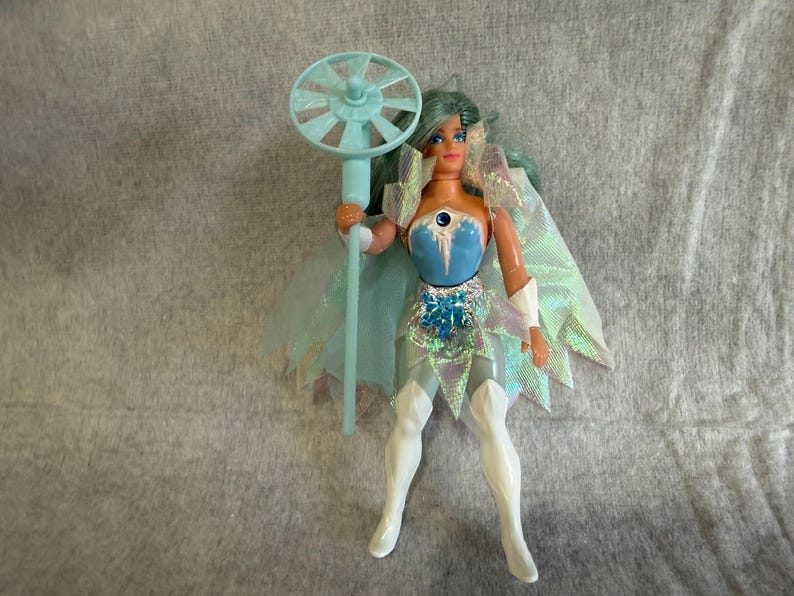 May include: A doll with teal hair wearing a blue and white bodice, a silver waist accent, and white boots. The doll features a translucent iridescent cape and holds a light blue staff topped with a circular, fan-like accessory. The figure has painted facial features and articulated limbs.