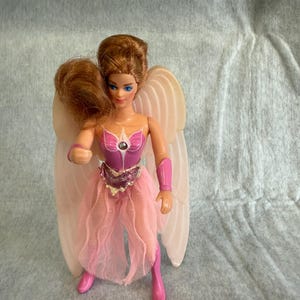 May include: A doll with auburn hair and blue eyes featuring large, pale, ribbed wings. The doll wears a pink bodice with a central jewel, a pink tulle skirt, pink arm cuffs, and pink boots. The figure has a fair complexion and a molded pose.