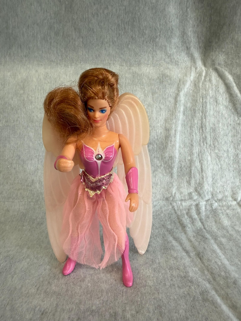 May include: A doll with auburn hair and blue eyes featuring large, pale, ribbed wings. The doll wears a pink bodice with a central jewel, a pink tulle skirt, pink arm cuffs, and pink boots. The figure has a fair complexion and a molded pose.