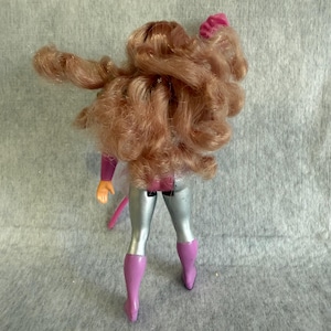 May include: A doll with long, wavy, light brown hair viewed from the back. The doll wears a purple and silver outfit consisting of a purple bodice, silver leggings, and purple boots. One arm is raised, and the other is positioned at the side.