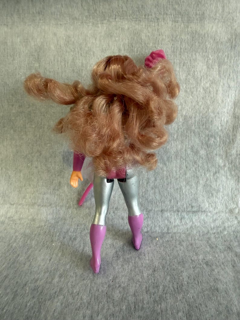 May include: A doll with long, wavy, light brown hair viewed from the back. The doll wears a purple and silver outfit consisting of a purple bodice, silver leggings, and purple boots. One arm is raised, and the other is positioned at the side.