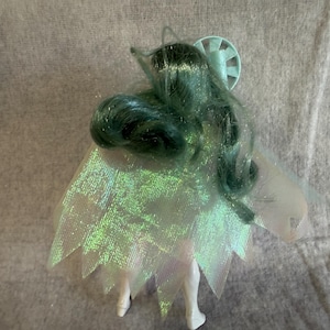 May include: A doll with long, wavy, dark teal hair and a light blue circular accessory on the head. The doll wears a translucent, iridescent cape with a jagged, pointed hemline that shimmers with green and purple tones over a white body.