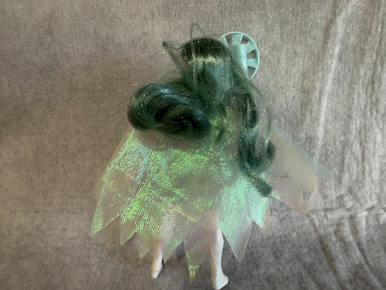 May include: A doll with long, wavy, dark teal hair and a light blue circular accessory on the head. The doll wears a translucent, iridescent cape with a jagged, pointed hemline that shimmers with green and purple tones over a white body.