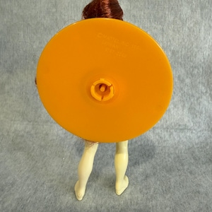 May include: A doll with reddish-brown hair featuring a large, circular, orange plastic base attached to its back. The base has a central connector and is embossed with the text Mattel Inc 1966, Taiwan, 9187-2049. The doll has pale legs.