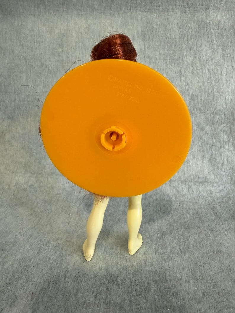 May include: A doll with reddish-brown hair featuring a large, circular, orange plastic base attached to its back. The base has a central connector and is embossed with the text Mattel Inc 1966, Taiwan, 9187-2049. The doll has pale legs.