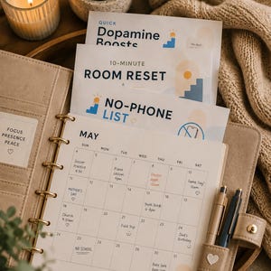 Può includere: Un'agenda ad anelli beige aperta su una pagina del calendario di maggio con note scritte a mano, accompagnata da tre schede impilate etichettate Quick Dopamine Boosts, 10-Minute Room Reset e No-Phone List, con accenti grafici blu e arancioni.