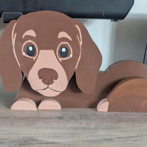 May include: A brown wooden cutout of a dachshund dog with floppy ears, a lighter tan muzzle, and large dark eyes with white highlights. The dog is depicted in a lying position with its front paws extended forward and its hindquarters curved.