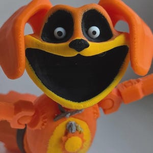 May include: A 3D printed orange dog figurine with a wide black mouth, large white eyes, and black eye patches. The figure features yellow accents on the face and chest, a small sun-shaped emblem on the torso, and articulated limbs.