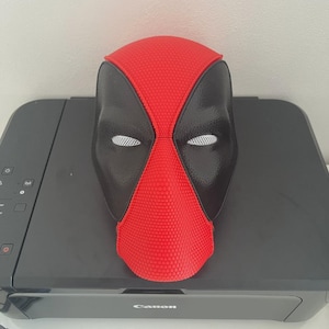 May include: A full-face mask featuring a vibrant red center panel with a textured, dotted surface and smooth black side panels. The mask includes white mesh eye coverings and a distinct seam running vertically down the center of the forehead and nose.