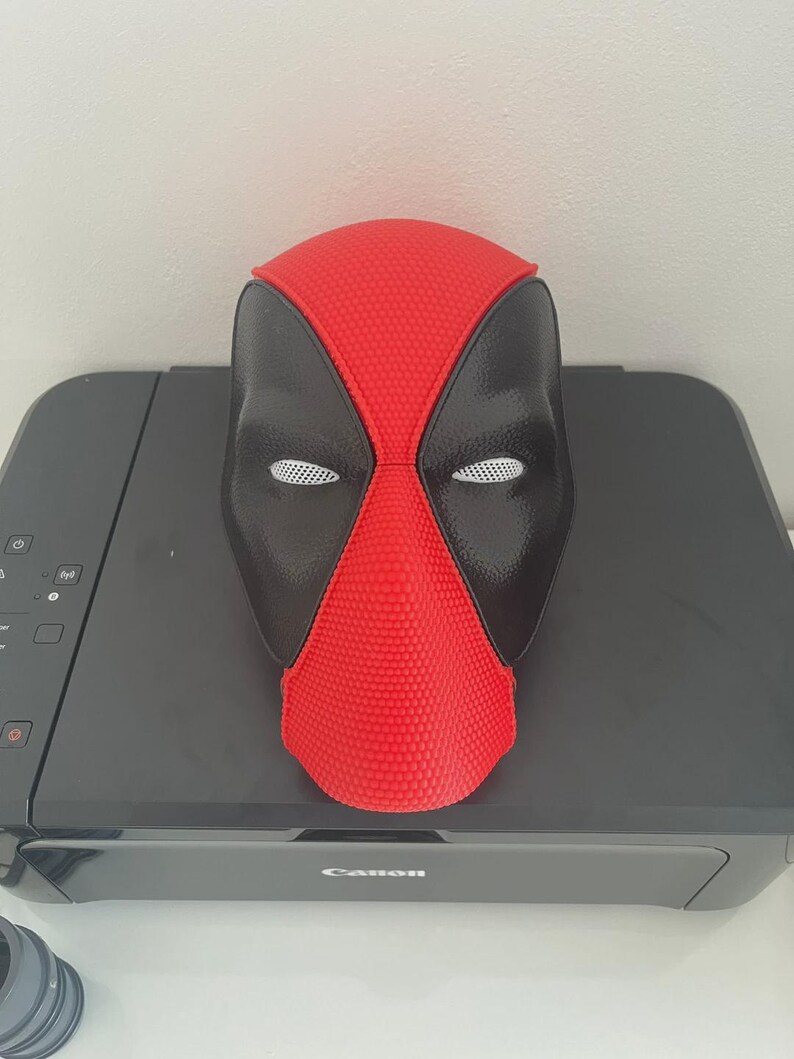 May include: A full-face mask featuring a vibrant red center panel with a textured, dotted surface and smooth black side panels. The mask includes white mesh eye coverings and a distinct seam running vertically down the center of the forehead and nose.