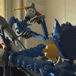 May include: A 3D-printed articulated action figure featuring a jester-like head with a white skull face, yellow and red accents, and a blue body. The figure has a mechanical appearance with visible joints, textured surfaces, and a sharp, pointed arm.