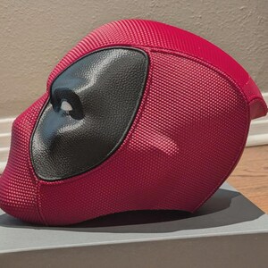 May include: A red full-face helmet featuring a textured, grid-like surface pattern and a smooth, black, almond-shaped eye piece. The helmet has a rounded, aerodynamic profile with a distinct chin section and a solid, dark red color scheme.