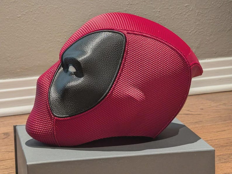 May include: A red full-face helmet featuring a textured, grid-like surface pattern and a smooth, black, almond-shaped eye piece. The helmet has a rounded, aerodynamic profile with a distinct chin section and a solid, dark red color scheme.