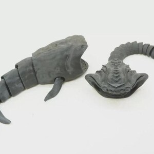 Puede incluir: Dos figuras articuladas de color gris oscuro, una con forma de ballena con cuerpo segmentado y boca abierta, y la otra con una cabeza ancha y plana y una cola larga, segmentada y curvada, ambas con un acabado de superficie mate y texturizado.