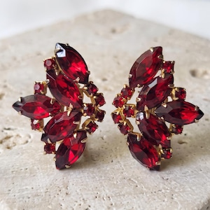 May include: A pair of gold-tone earrings featuring a cluster of deep red marquise and round-cut faceted crystals. The stones are arranged in a symmetrical, leaf-like pattern, secured in metal settings that highlight the vibrant red color.