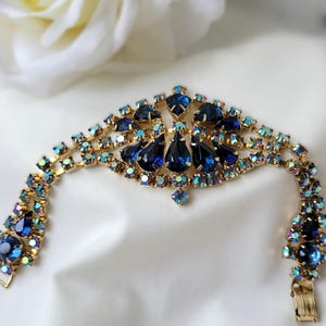 May include: A gold-tone bracelet featuring a central cluster of deep blue pear-shaped rhinestones surrounded by smaller iridescent crystals. The band consists of multiple rows of shimmering light-catching stones set in a metallic frame with a clasp.