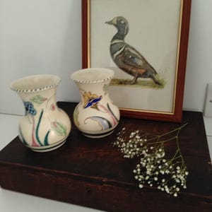 May include: Two cream-colored ceramic vases with scalloped rims and hand-painted floral motifs in blue, green, and yellow. Behind the vases is a framed watercolor painting of a duck in a brown wooden frame, accompanied by a sprig of baby's breath.