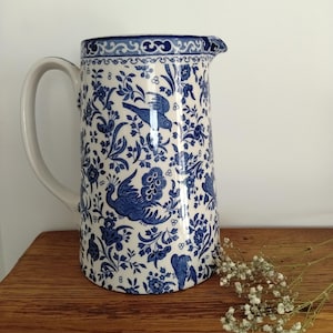 Puede incluir: Una jarra de cerámica con base blanca y un patrón azul de flores y aves. El diseño incluye pavos reales estilizados, pájaros pequeños y follaje intrincado. Un borde decorativo recorre la parte superior y un asa curva está fijada en el lateral.