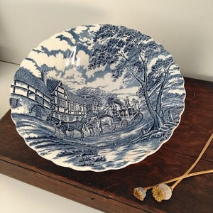 May include: A round ceramic bowl with a scalloped edge featuring a blue and white transferware pattern. The design depicts a horse-drawn carriage travelling past timber-framed buildings and trees under a cloudy sky, rendered in a classic style.