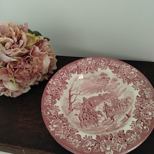 May include: A round ceramic plate featuring a pink and white transferware pattern. The centre depicts a historical scene with a horse-drawn carriage and figures, while the wide rim is decorated with a detailed floral and scrollwork border.